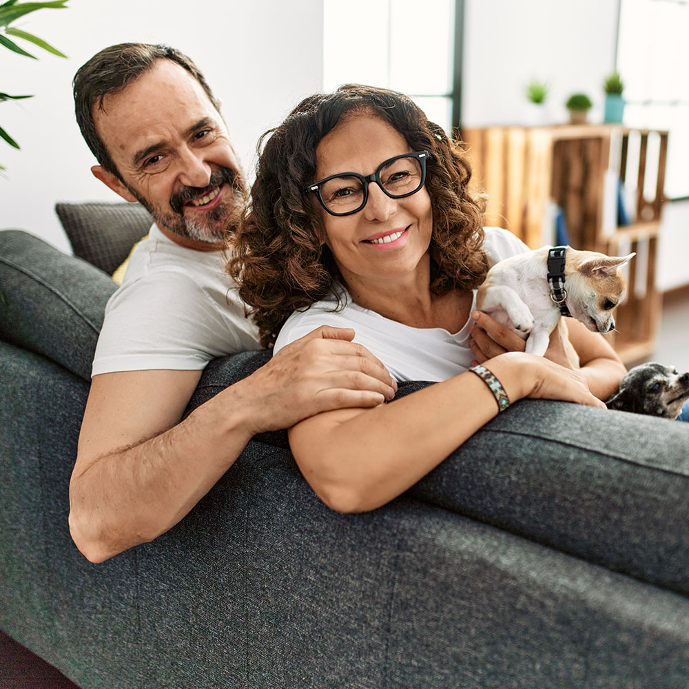 Middle aged couple with 2 small dogs sitting on a sofa, looking back at the camera.